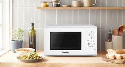 Frontansicht der Mikrowelle NN-E20JWMEBG von Panasonic mit geschlossener Tür auf einer Küchenarbeitsplatte. Rundherum befinden sich Lifestyle-Elemente wie eine Flasche Olivenöl, eine Schüssel mit ungekochter Pasta und ein Holzlöffel, die eine gemütliche Kochatmosphäre erzeugen.