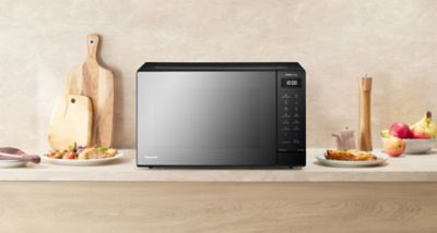 Panasonic GT65 microwave with black mirror finish on a kitchen counter, with cutting boards, a pepper mill, fruits, and plates of food in the background.