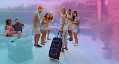 Pessoas reunidas ao ar livre a desfrutar de uma festa enquanto alguém puxa uma coluna de som portátil Panasonic SC‑BMAX30 com anéis de LED iluminados.