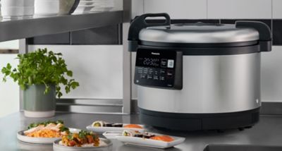 Un cuiseur à riz Panasonic Pro IH est posé sur le comptoir d'une cuisine, avec des assiettes de rouleaux de sushi et des plats de riz placés devant lui, à côté d'une plante en pot.