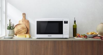 Bela mikrovalovna pečica Panasonic CT55 stoji na kuhinjskem pultu, okoli nje pa so razporejene deske za rezanje, kruh, steklenica olivnega olja in krožnik hrane.