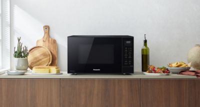 Cuptorul cu microunde Panasonic CT56, de culoare neagră, este așezat pe blatul de bucătărie, înconjurat de tocătoare, pâine, o sticlă de ulei de măsline și o farfurie cu mâncare.