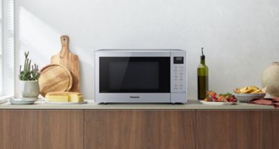 Cuptorul cu microunde Panasonic CT57, de culoare argintie, este așezat pe blatul de bucătărie, înconjurat de tăvi de tăiat, pâine, o sticlă de ulei de măsline și o farfurie cu mâncare.