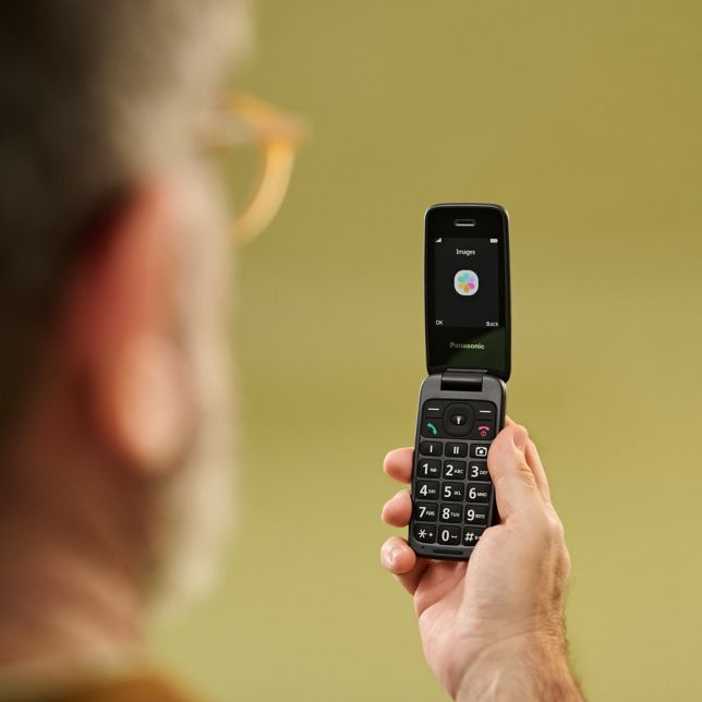 Zbliżenie dłoni trzymającej telefon Panasonic K‌X-T‌U410, na którym widać duży kolorowy ekran i dużą klawiaturę na czerwonym