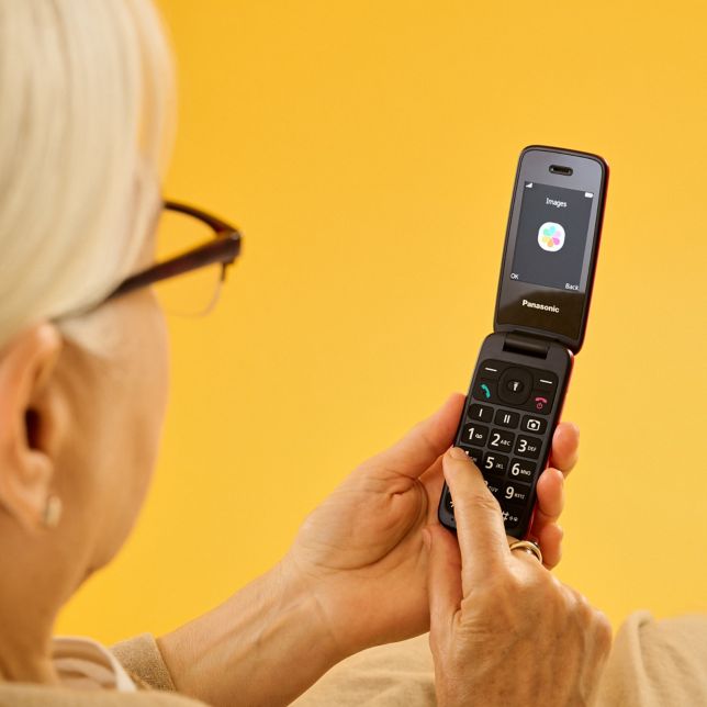 Kobieta w okularach trzyma w ręku otwarty telefon z klapką Panasonic K‌X-T‌U410 na żółtym tle.