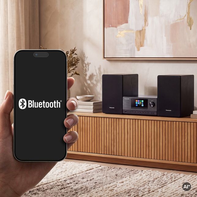 Egy kéz, amely egy Bluetooth logót mutató okostelefont tart egy fa tálalószekrényen álló Panasonic SC-PMX112 előtt, a vezeték nélküli párosítást szemléltetve.