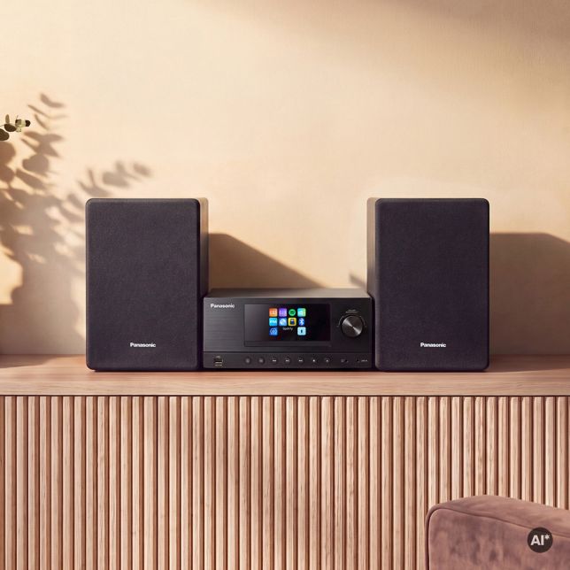A Panasonic SC-PMX112 faházas hangszórói egy bordázott tálalószekrényen, egy szárított lombozattal teli váza mellett helyezkednek el, kiemelve a minimalista dizájnt.